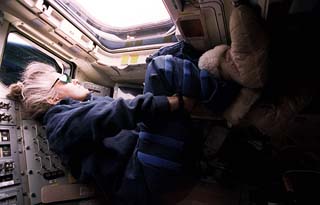 Lucid, wearing blue warm-ups, sun glasses and soft boots floats in the sunlight of the Atlantis aft flight deck window. 